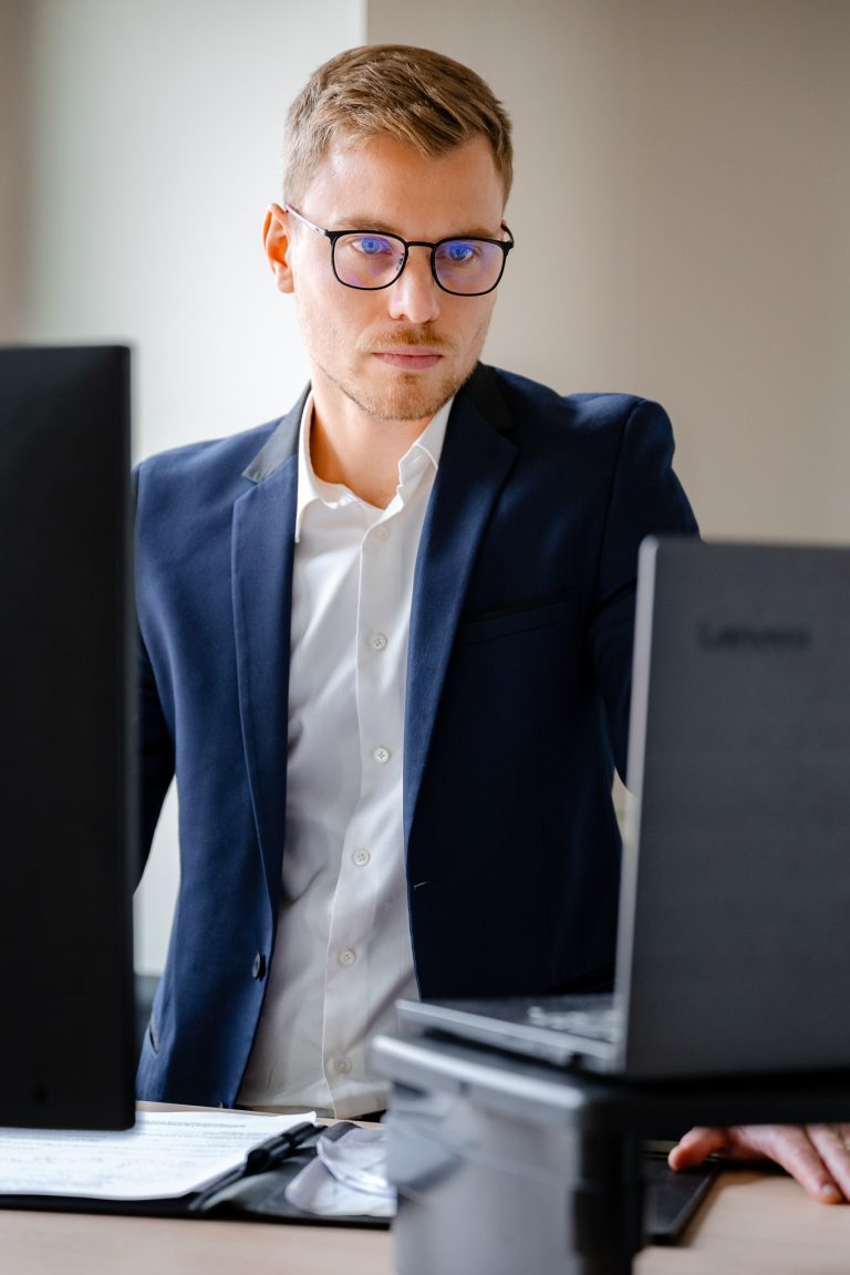 Reportage photo d'entreprise Lyon : jeune professionnel en costume bleu marine et lunettes concentré devant son double écran d'ordinateur au bureau, photographe corporate spécialisé dans les portraits professionnels authentiques